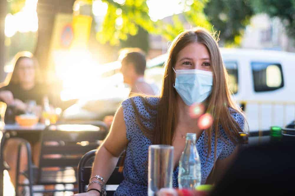 woman sitting at table wearing a mask waiting for food with others spaced apart in the background at an outdoor party during covid