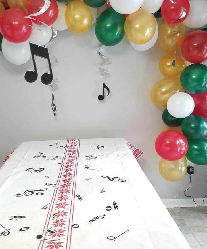 green, red, white and gold balloon garland with music notes hung over table with white linen and musical notes on table