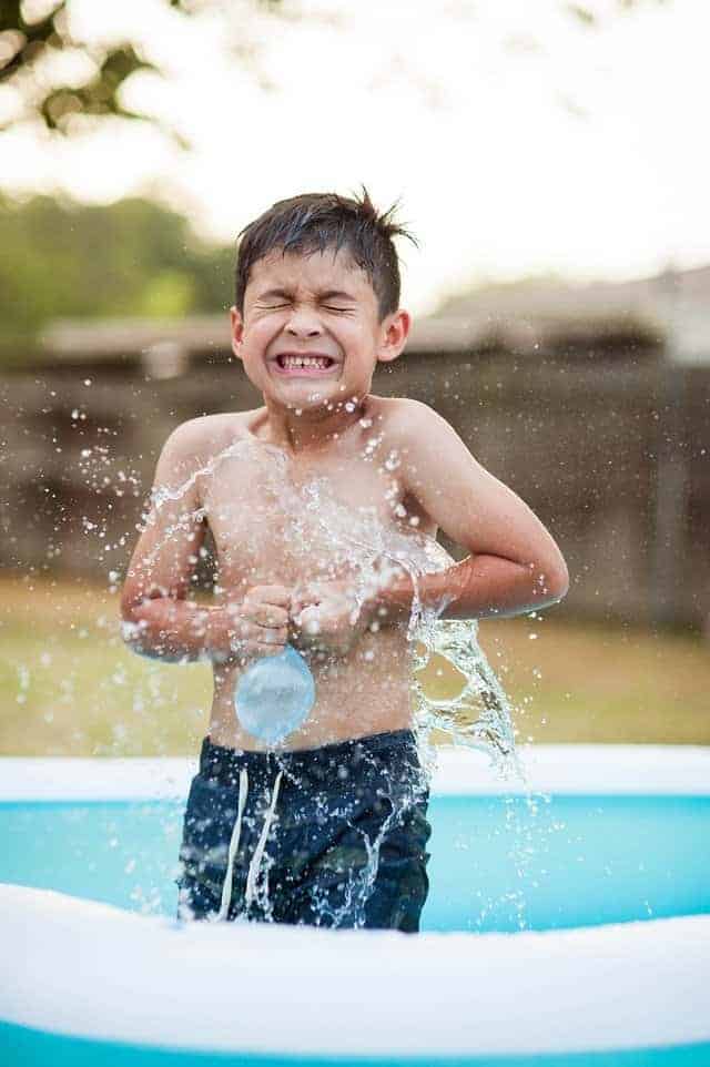 boy wincing as water balloon splashes on him in a small pool