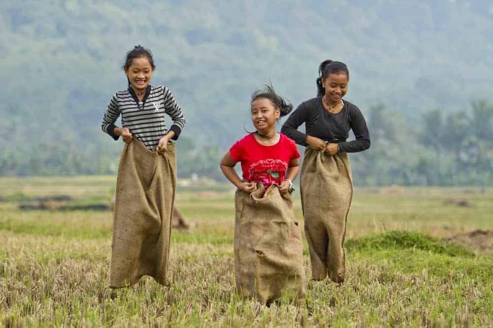 three girls laughing during a potato sack race in field with mountains and trees in background