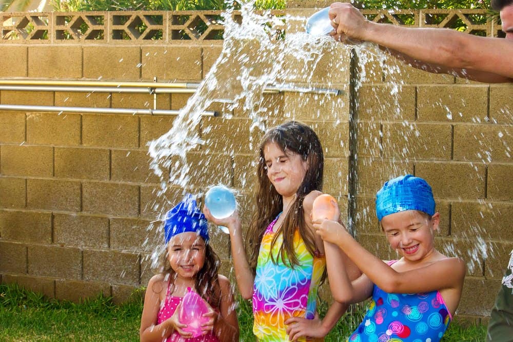 The arms of a father break a blue water balloon over the heads of his three daughters.  The girls are wearing bathing suits and two of them have swim caps on.