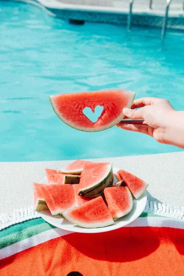 hand holding slice of watermelon with heart cutout over a plate of watermelon slices by pool