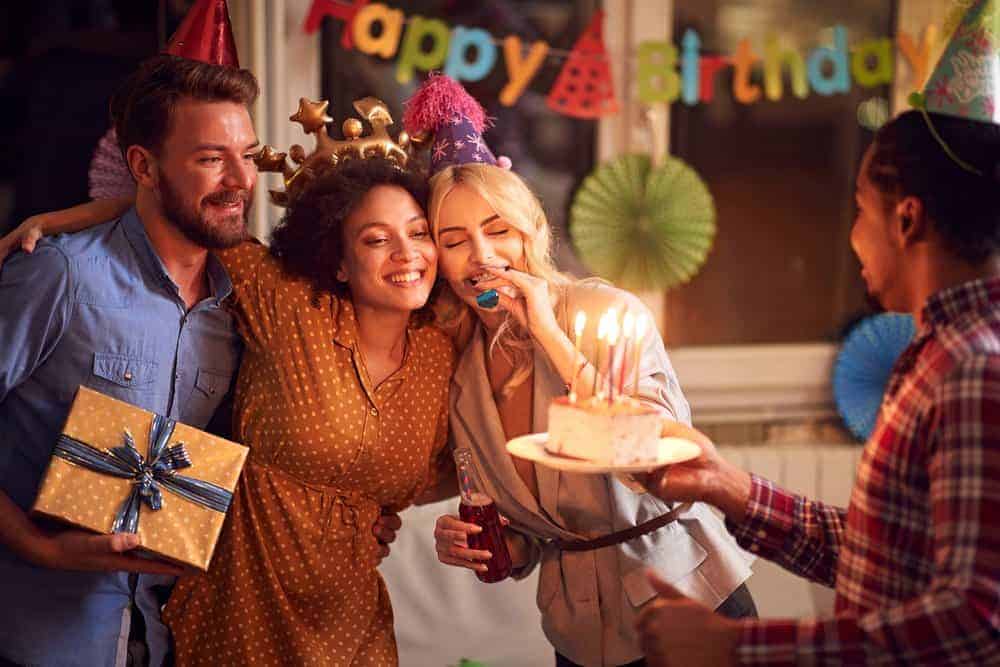 Adults celebrating a friend's birthday party with party decoration, a gift and cake with lit candles