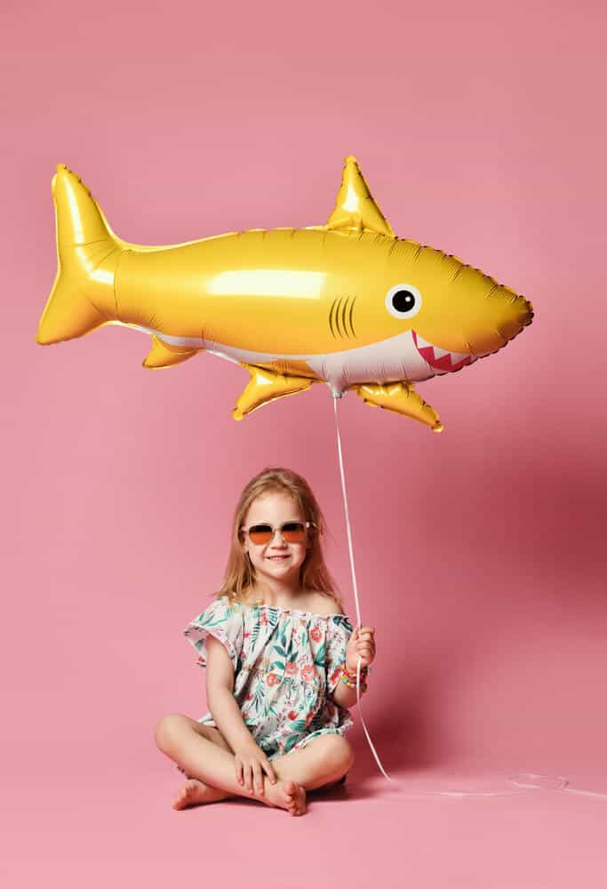 girl sitting on pink background holding yellow shark party decoration balloon