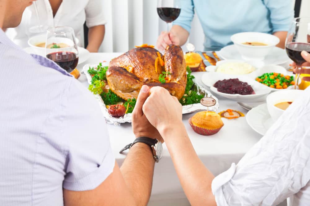 individuals holding hands praying with thanksgiving turkey on table behind hands