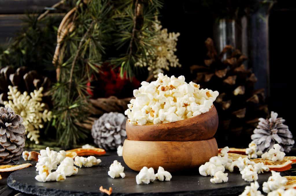 popcorn in wooden bowl with some spilling out in front of a rustic holiday background of evergreen branches and pinecones
