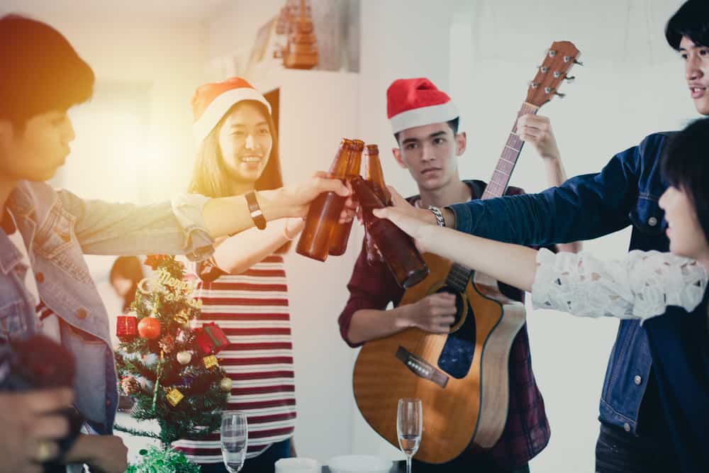 group of asian friends in santa hats toasting beer bottles with one man playing guitar