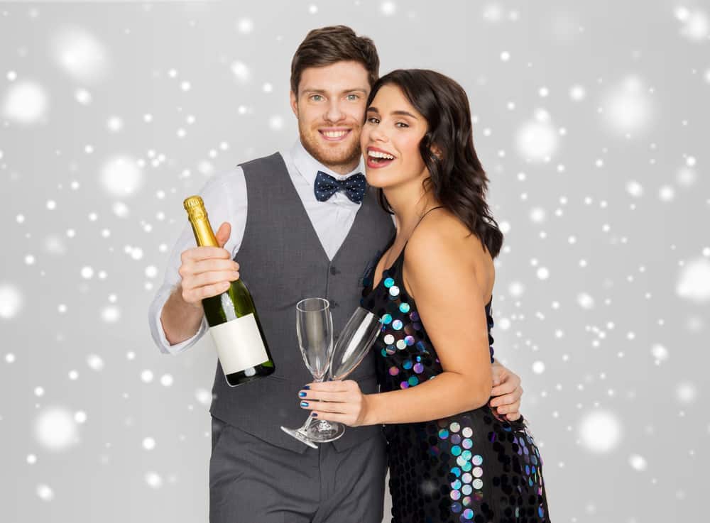 man and woman pose for a photo with champagne and glasses wearing fancy holiday attire with white sparkly background