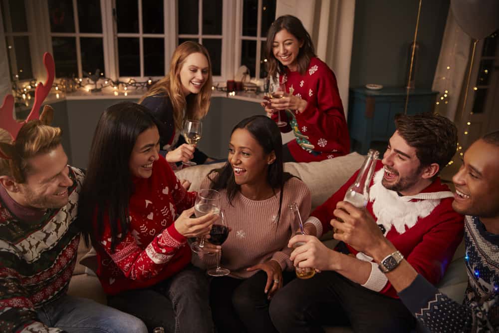 group of inter-racial friends sitting on couch in holiday sweaters laughing while drinking alcoholic beverages