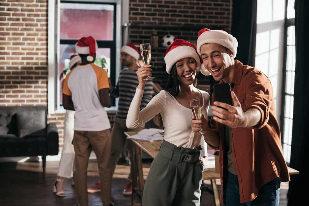 man and woman take selfie at the workplace while wearing santa hats and drinking champagne with coworkers in background