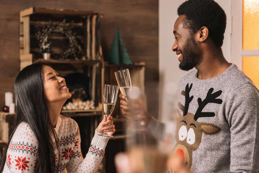 asian woman and black man cheers champagne glasses while wearing nordic holiday sweaters with a scandinvian vibe background of rustic decor