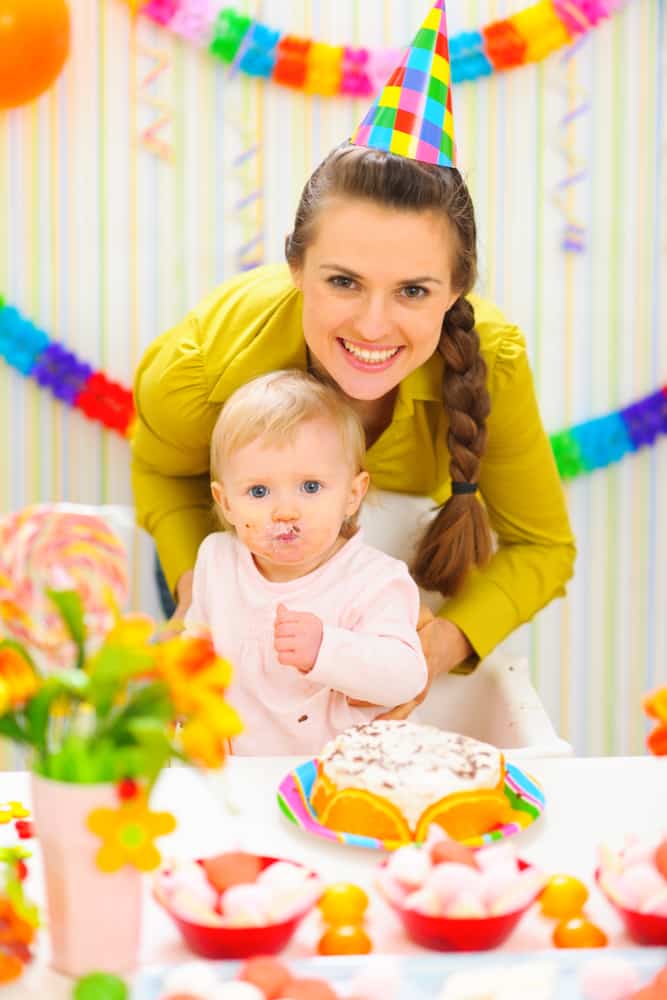mother and baby at baby's first birthday party with table of cake, foods and first birthday party favors for 1 year olds