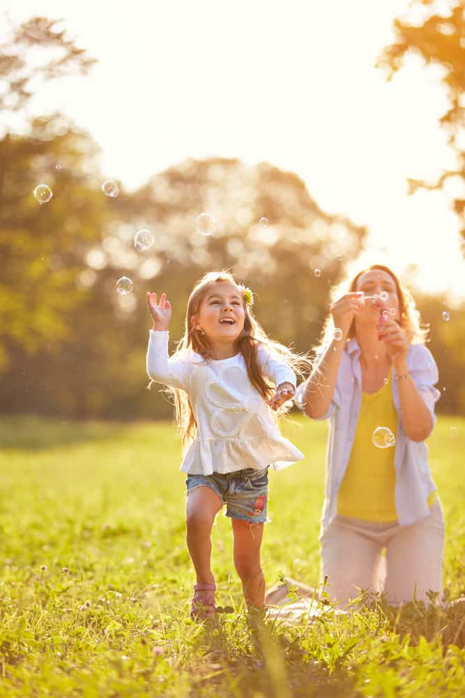 small child chasing bubbles as a mother blows them outside in the background