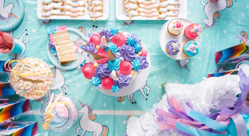 overhead view of a unicorn party table with unicorn tablecloth, unicorn cake and cupcakes and unicorn pinata