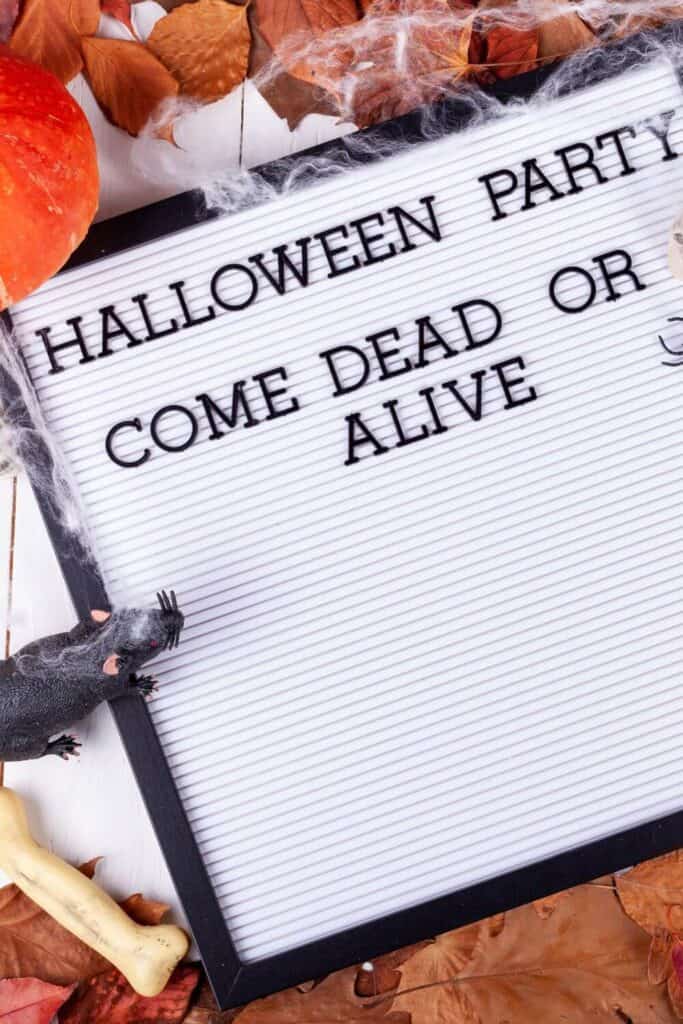 black letters on white letterboard that says halloween party come dead or alive with halloween spooky items surrounding
