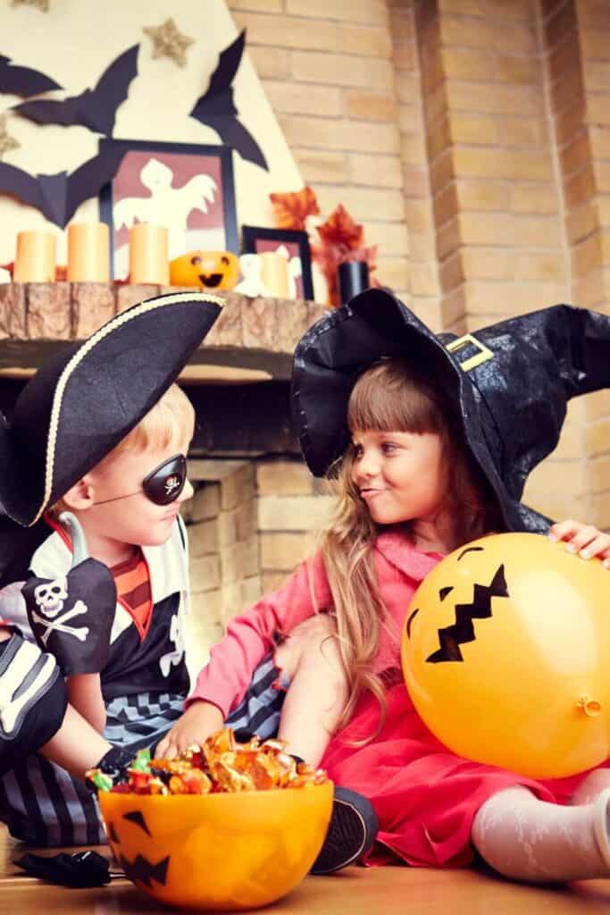 kids dressed up as a pirate an witch sitting on the floor at a halloween party grabbing candyf rom a pumpkin bowl