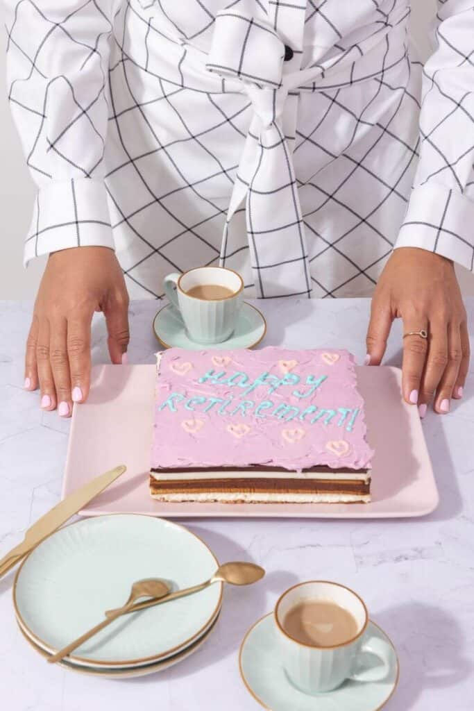 pink layered retirement cake with happy retirement write on it on a table with teacups and woman standng over it